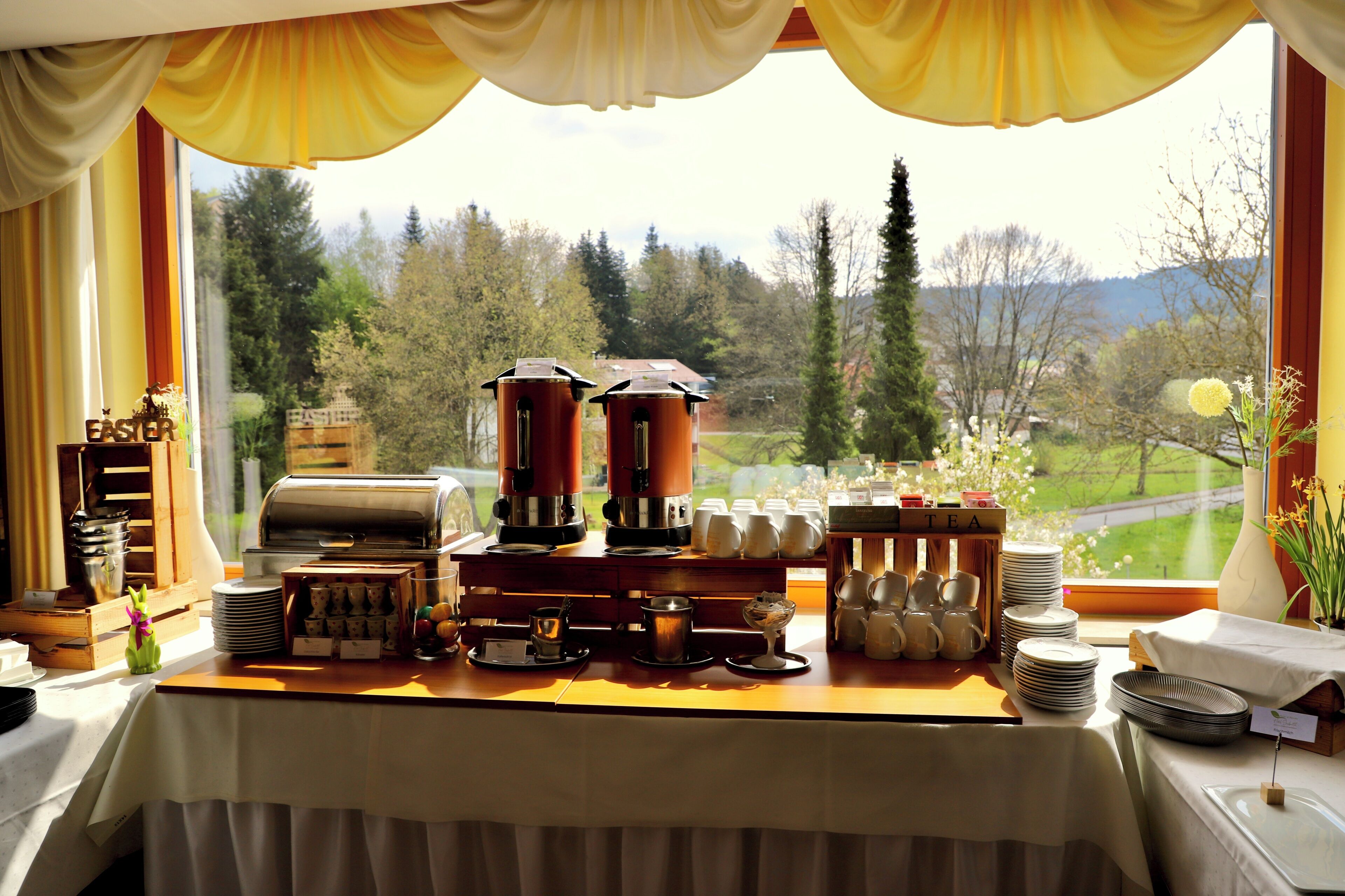 Montaje de un bufé de desayuno con máquinas de café, servicio de té y vistas a un paisaje verde.