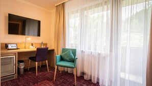 A hotel room with a desk, a chair, a television, and a window with sheer curtains.