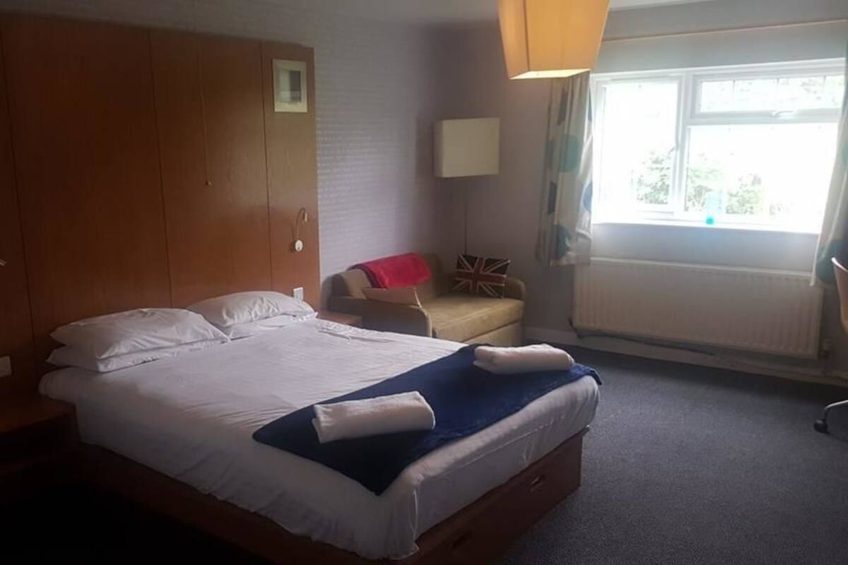 A neatly made bed with white linens and a blue blanket, a wooden wardrobe, a bedside table with a lamp, a small chair with a red cushion, and a window with curtains.