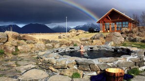 A rustic cabin with a stone hot tub in front, surrounded by large rocks and a mountain backdrop.