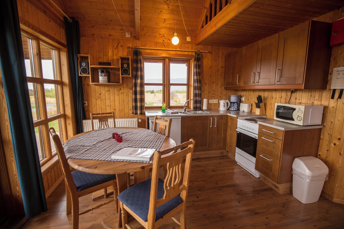A wooden cabin interior with a dining table, chairs, a kitchen with appliances, and a view through the window.