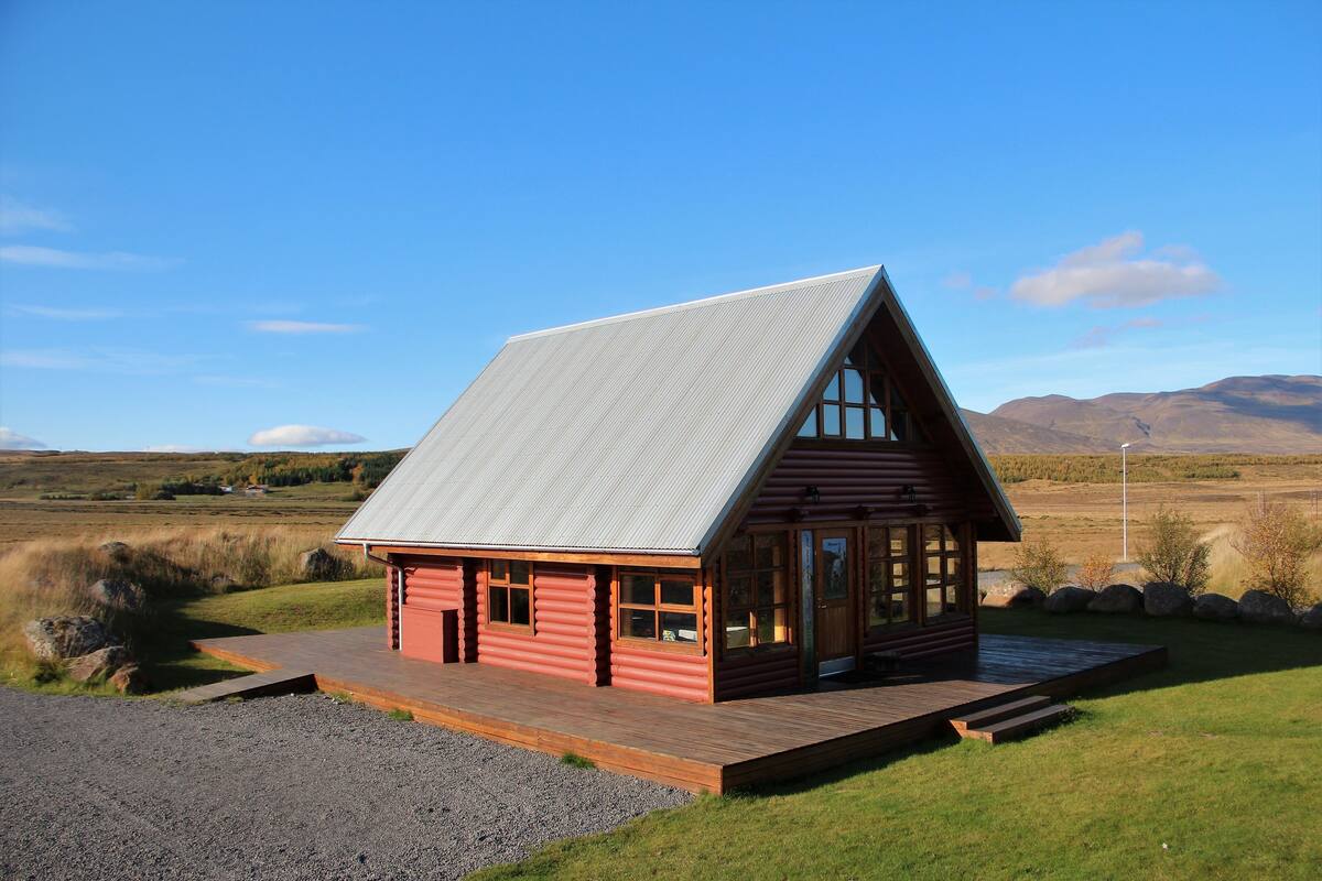 Una cabaña de troncos con techo metálico inclinado, terraza de madera y vista del paisaje circundante.