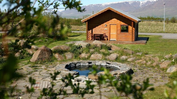 A wooden cabin with a hot tub, surrounded by rocks and greenery, with mountains in the background.