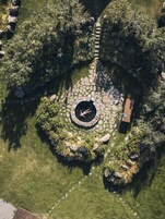 An aerial view of a circular stone pathway with a central fire pit, surrounded by lush greenery and a wooden bench.