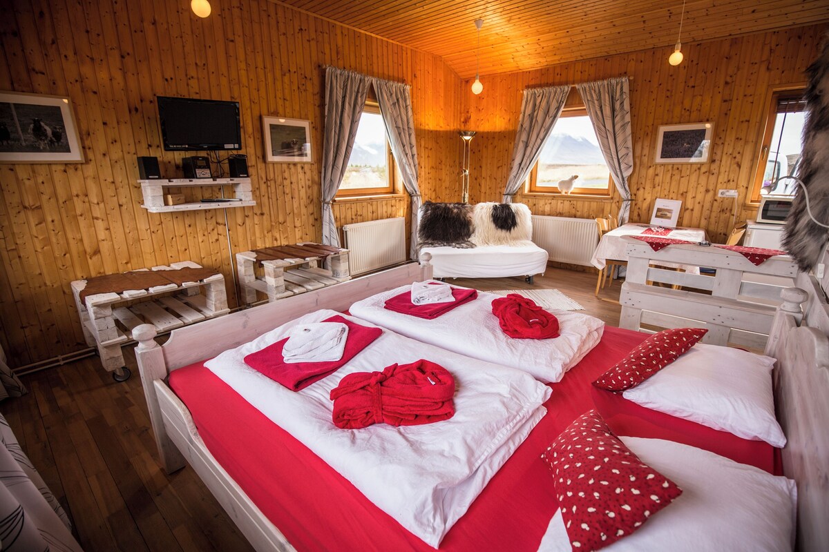 A wooden cabin room with a bed, a chair, a table, a television, and a window with curtains.