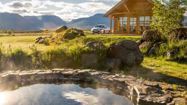 Une cabane en bois avec une voiture garée devant, un petit étang et des montagnes en arrière-plan.