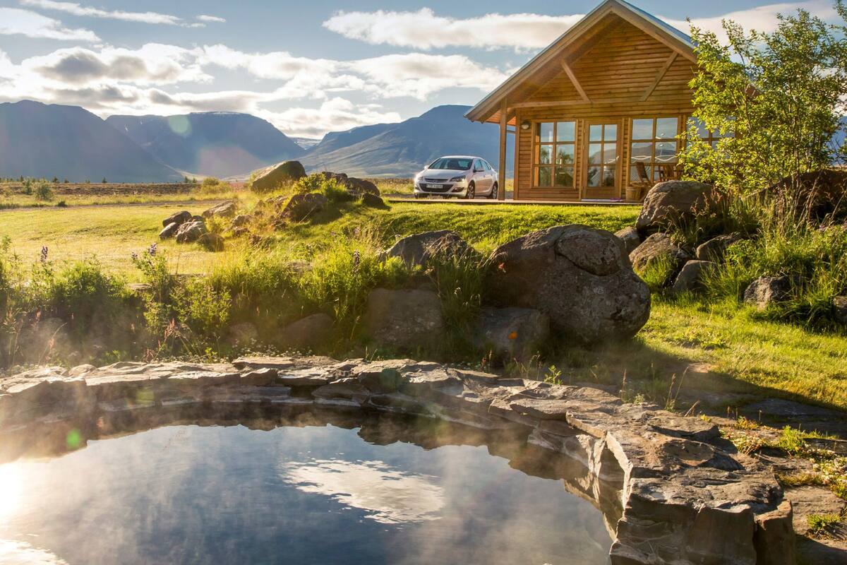 Una cabaña de madera con un coche estacionado adelante, un pequeño estanque y montañas al fondo.