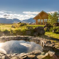 A wooden cabin with a car parked in front, a small pond, and mountains in the background.