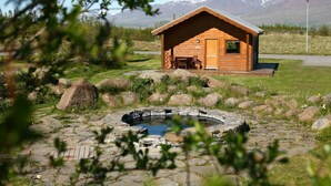 A wooden cabin with a hot tub and a dining area, surrounded by rocks and greenery, with mountains in the background.