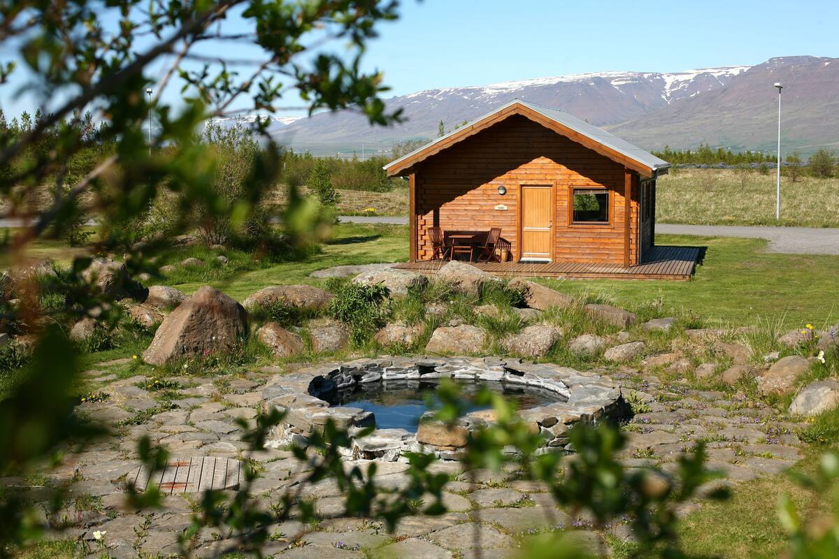 Una cabaña de madera con hidromasaje rodeada de rocas, un patio con sillas y vistas a la montaña al fondo.