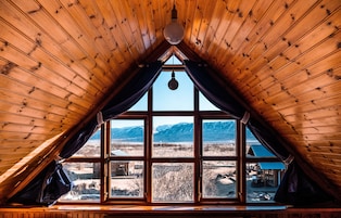 Una habitación con techo de madera inclinado y ventanas amplias que ofrecen vista a montañas y una cabaña.
