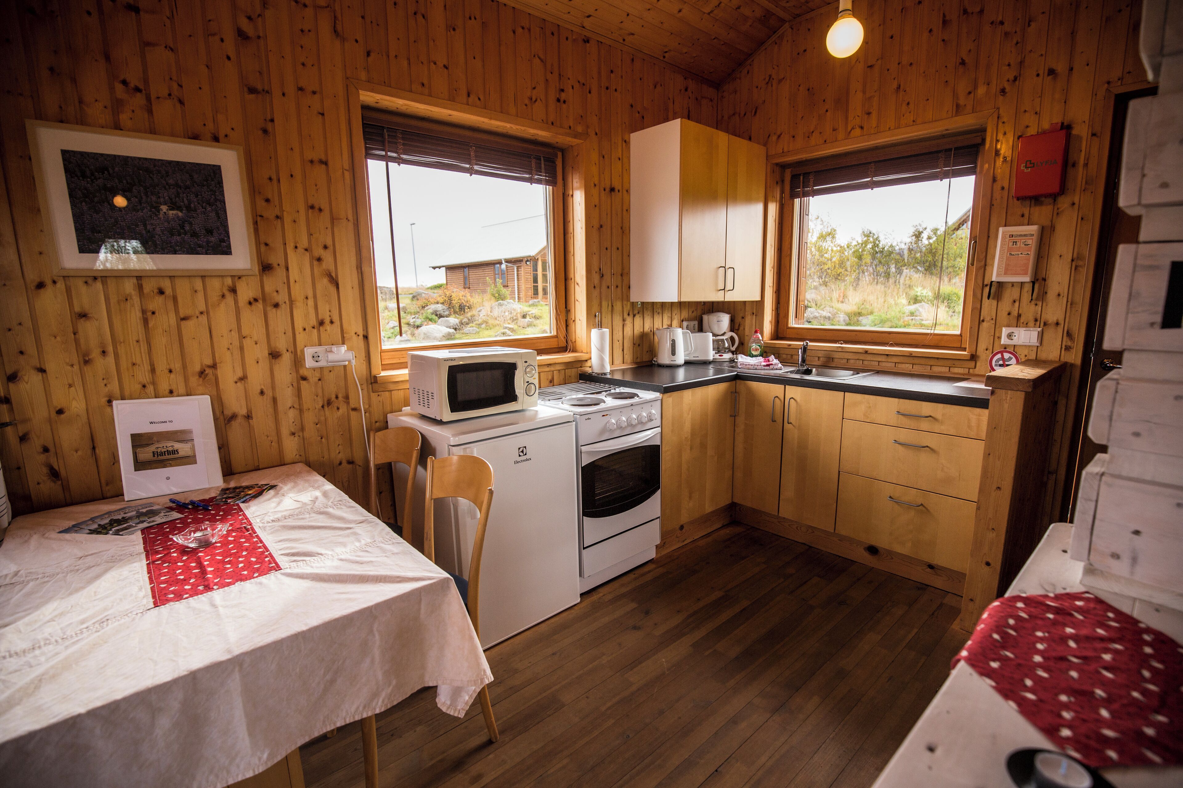 A compact kitchen with wooden walls, a microwave, a stove, and a sink. There is a table with a red placemat and a chair.