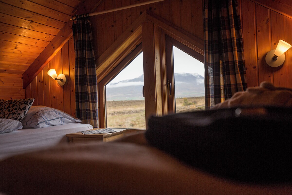 Habitación de una cabaña de madera con una cama, una mesa y vistas a un paisaje montañoso a través de un ventanal grande.
