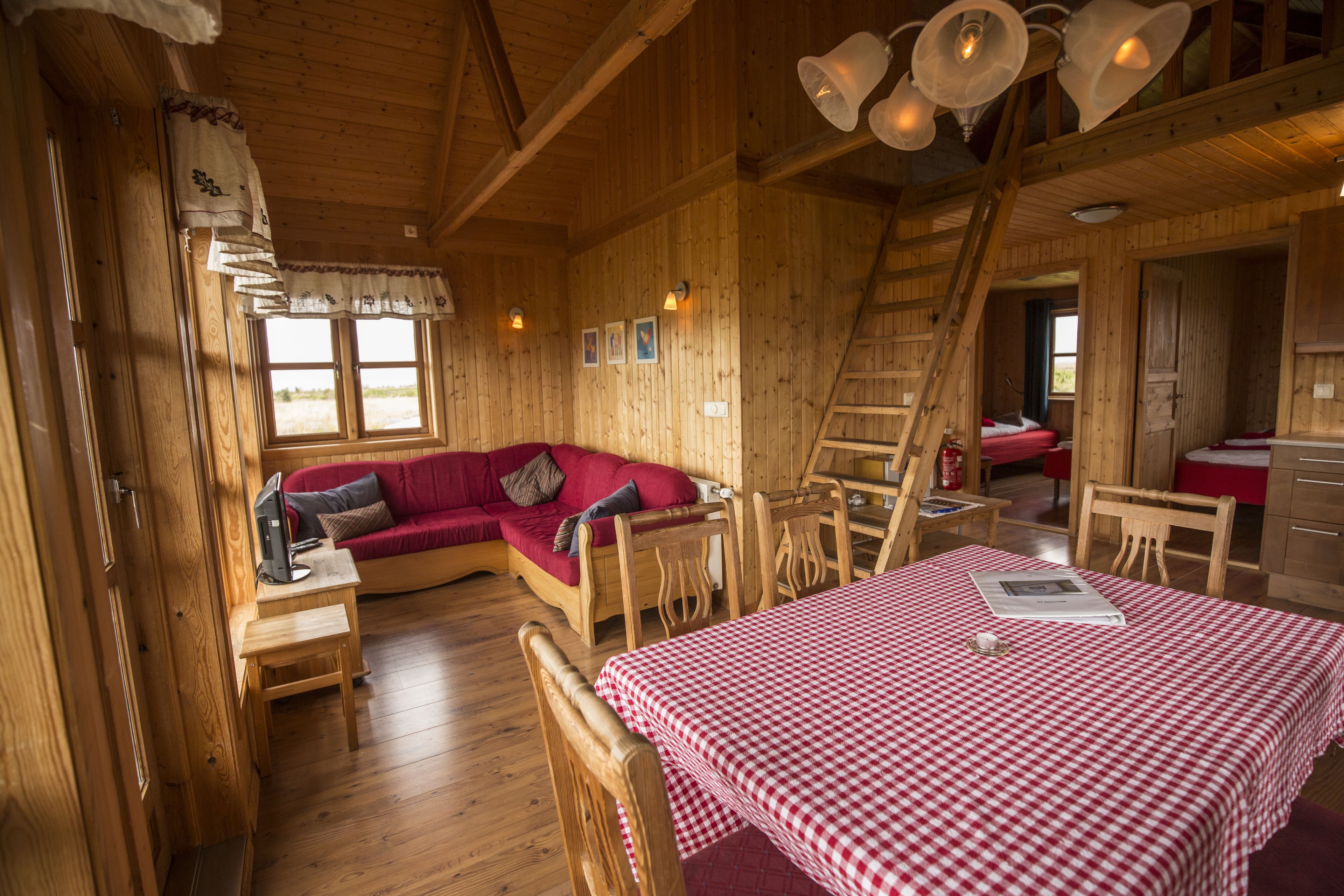 Interior de una cabaña de madera con una cama a cuadros roja y blanca, un sofá rojo, mesas de madera y una escalera.