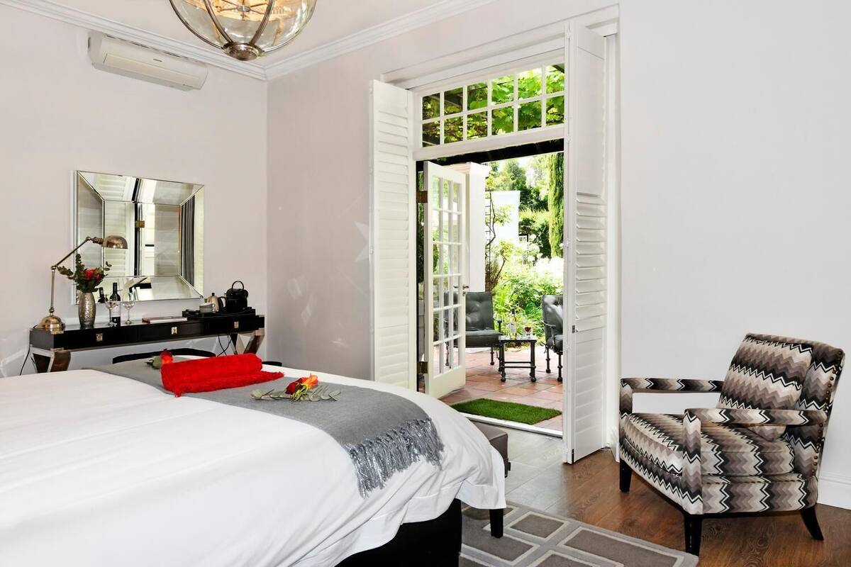 A hotel room with a bed, a desk with a mirror, a chair, and an open door leading to a patio with outdoor furniture.