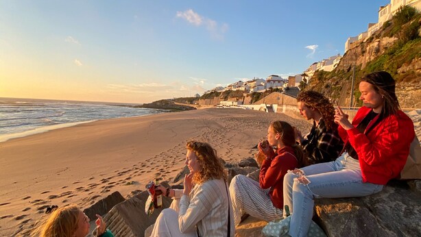 A group of people sitting on a stone wall by the beach, overlooking the ocean and a coastal town. - Surfer's Den Ericeira (Mafra)