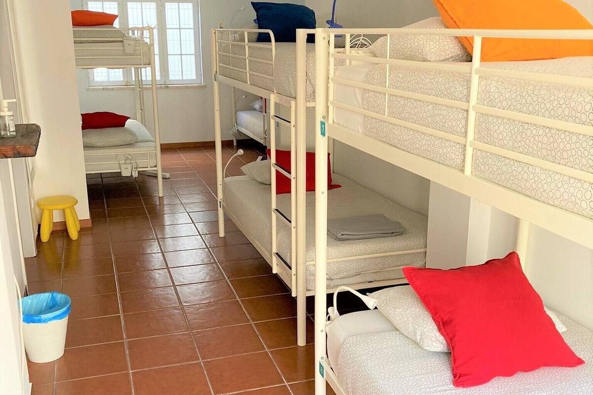 Een slaapzaal met stapelbedden, een klein krukje, een prullenbak en een raam met jaloezieën.