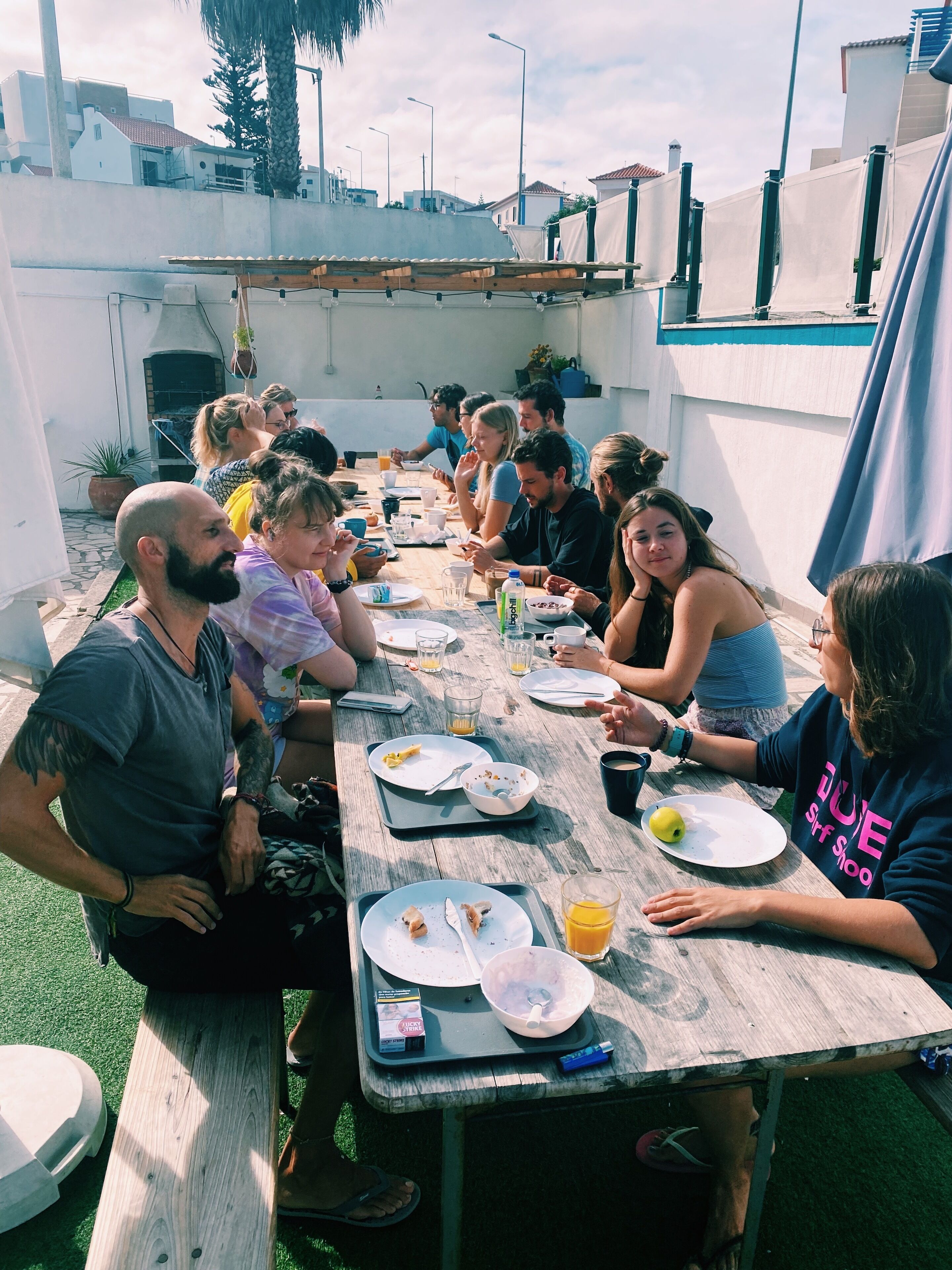 A group of people sitting around a wooden table outdoors, eating and drinking.