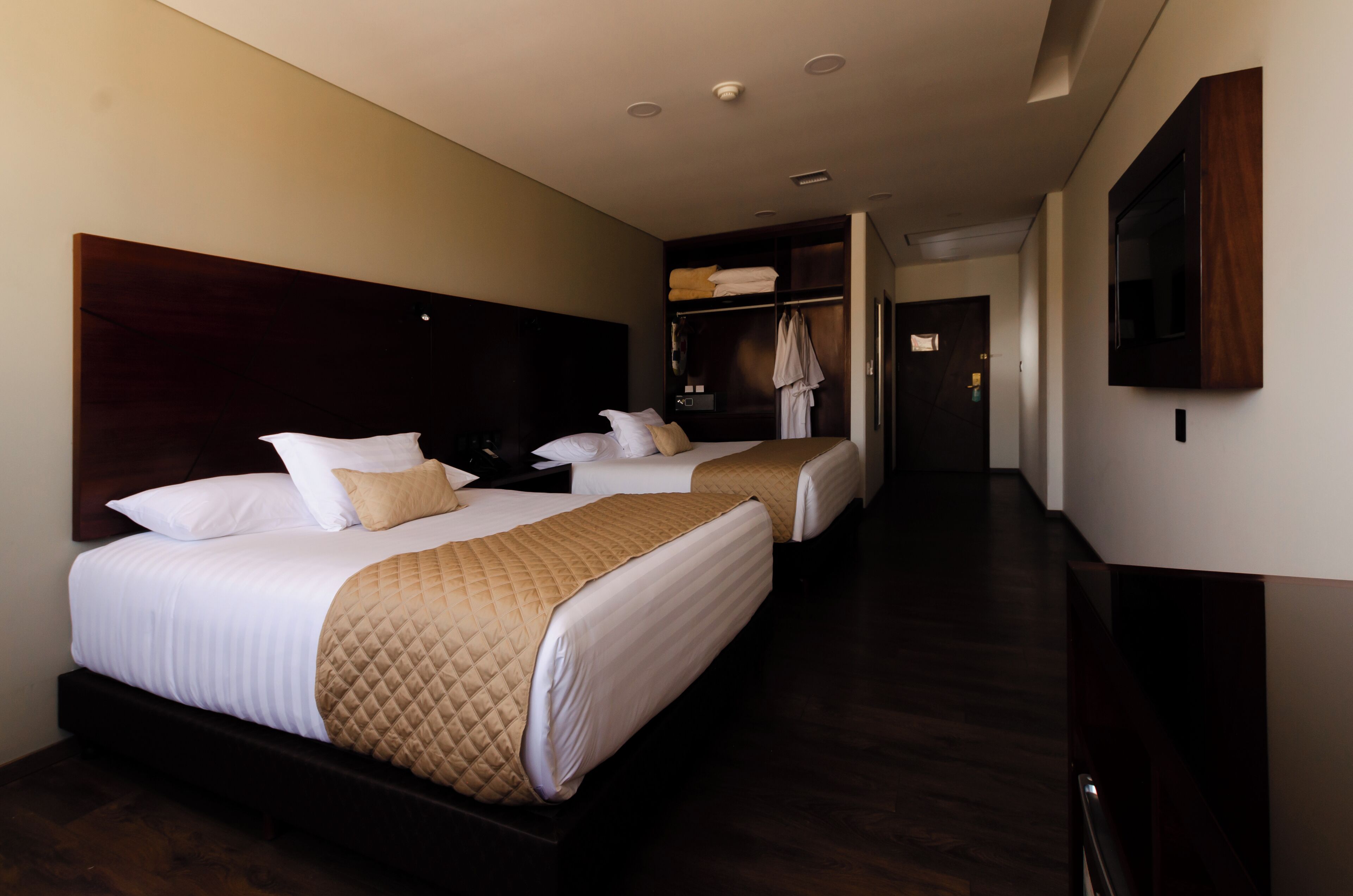 A hotel room with two beds, a dark wooden headboard, a wardrobe, and a flat-screen TV mounted on the wall.