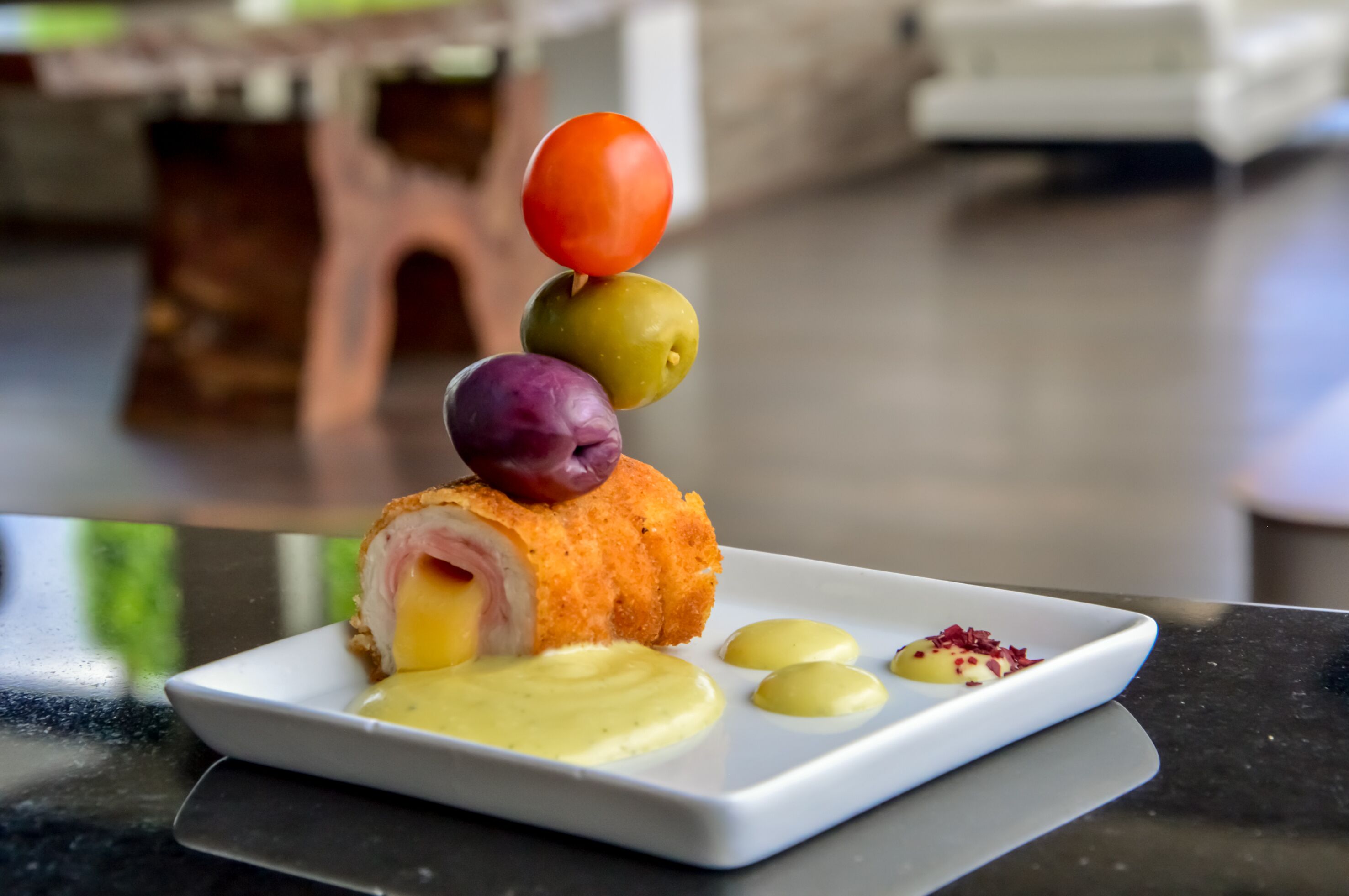Un plato con un rollo de carne y queso, acompañado de una brocheta de frutas y una pequeña porción de salsa.