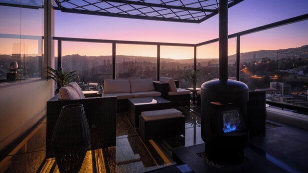 A modern rooftop lounge with a glass balustrade, comfortable seating, and a wood-burning stove.