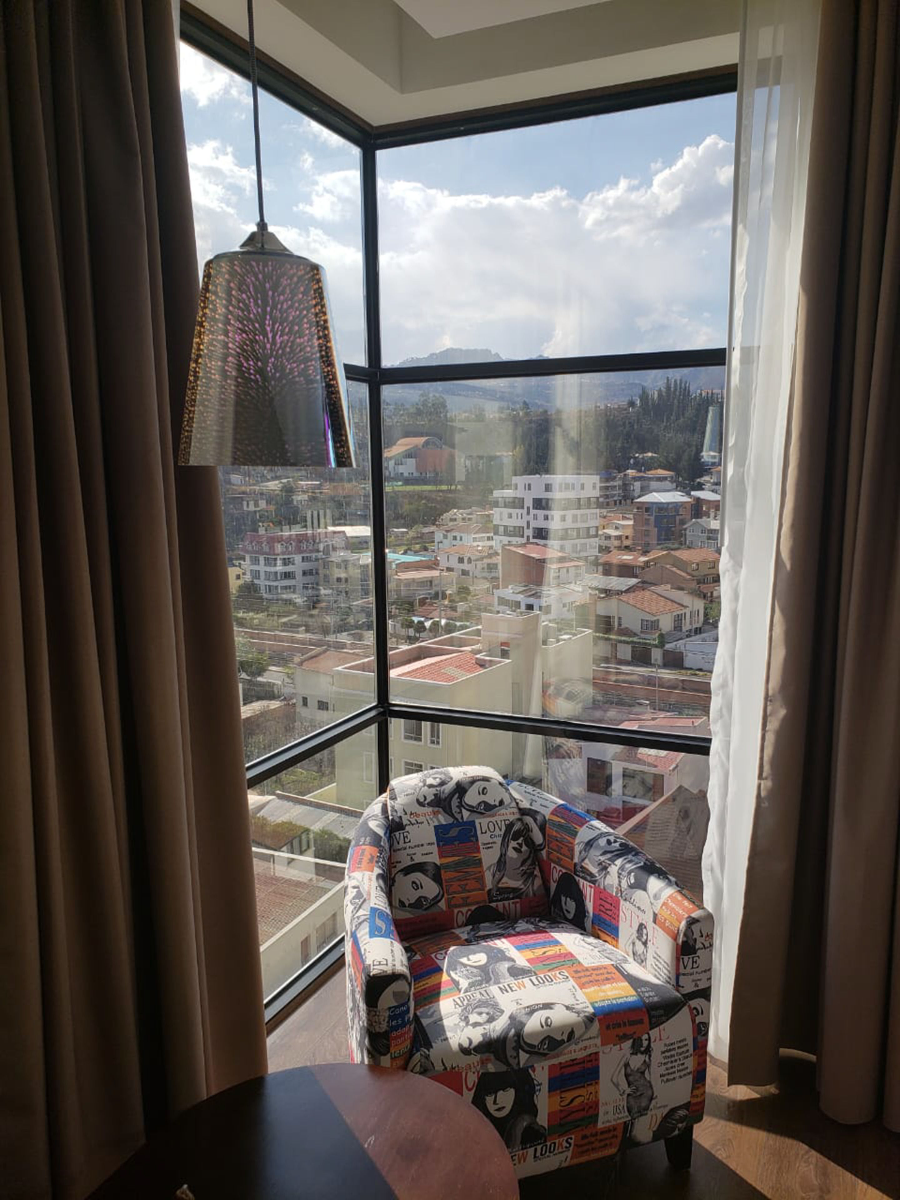 Habitación de esquina con un ventanal amplio que ofrece vistas a la ciudad, un sillón estampado y una lámpara colgante.