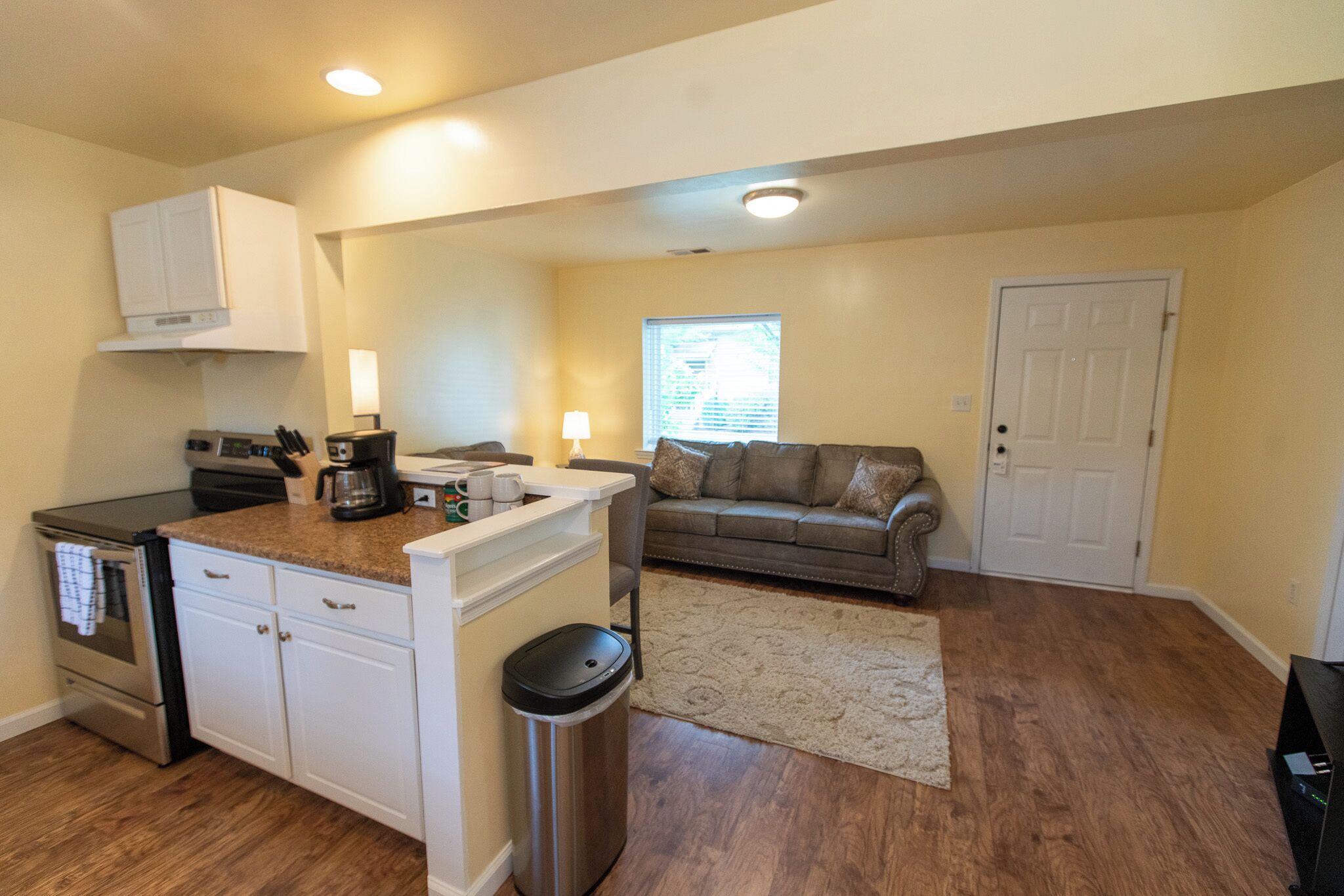 A compact kitchen with a stove, microwave, and coffee maker, a small island with a trash can, a gray sofa, and a white door.