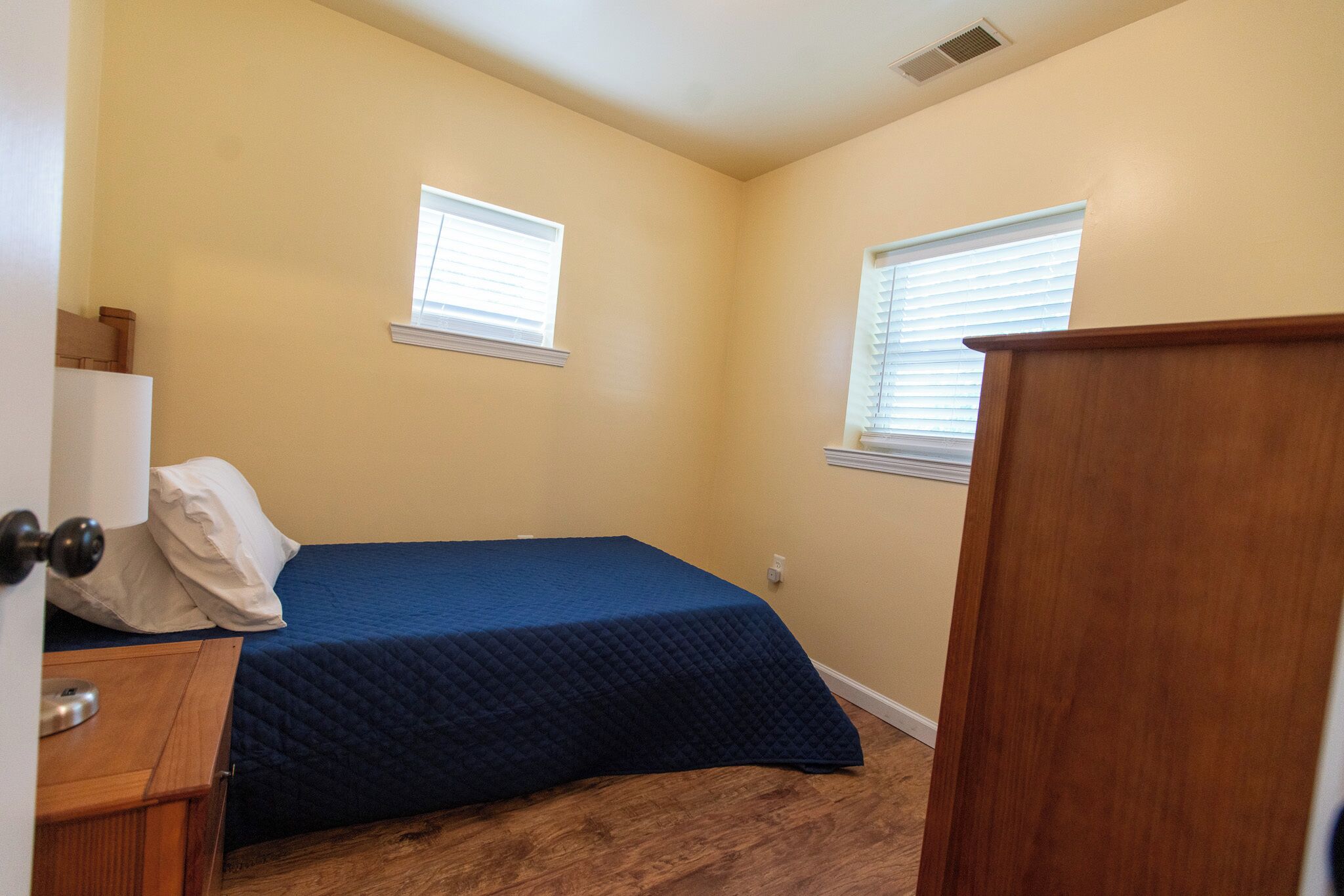 Un dormitorio con una cama, dos ventanas, un aparador de madera y una puerta.