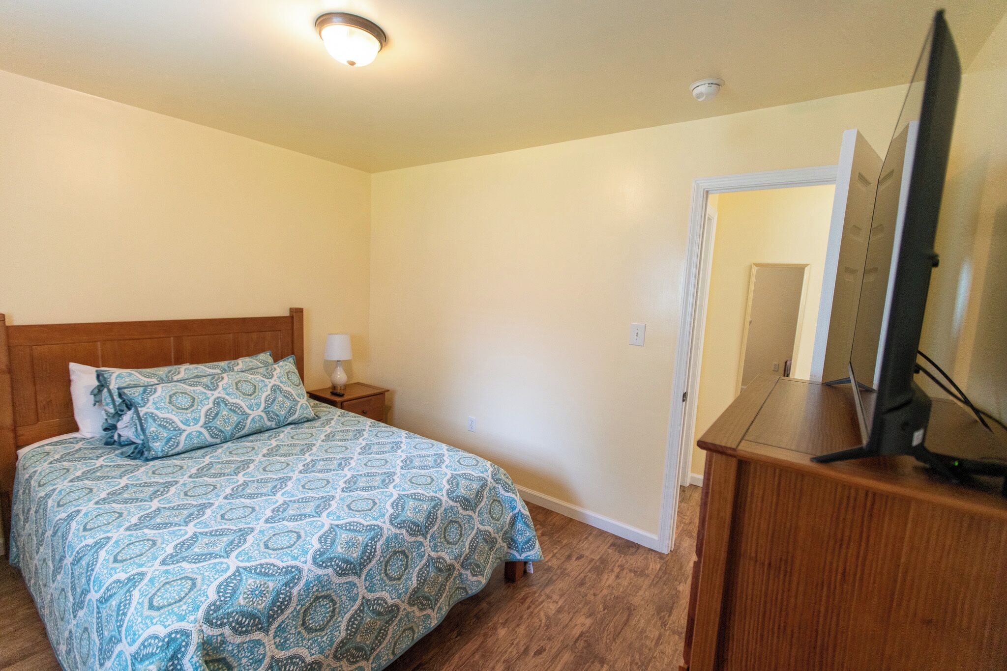 Un dormitorio con cabecera de madera, colcha estampada, mesita de noche con lámpara, cómoda de madera con televisor de pantalla plana y una puerta que da a otra habitación.