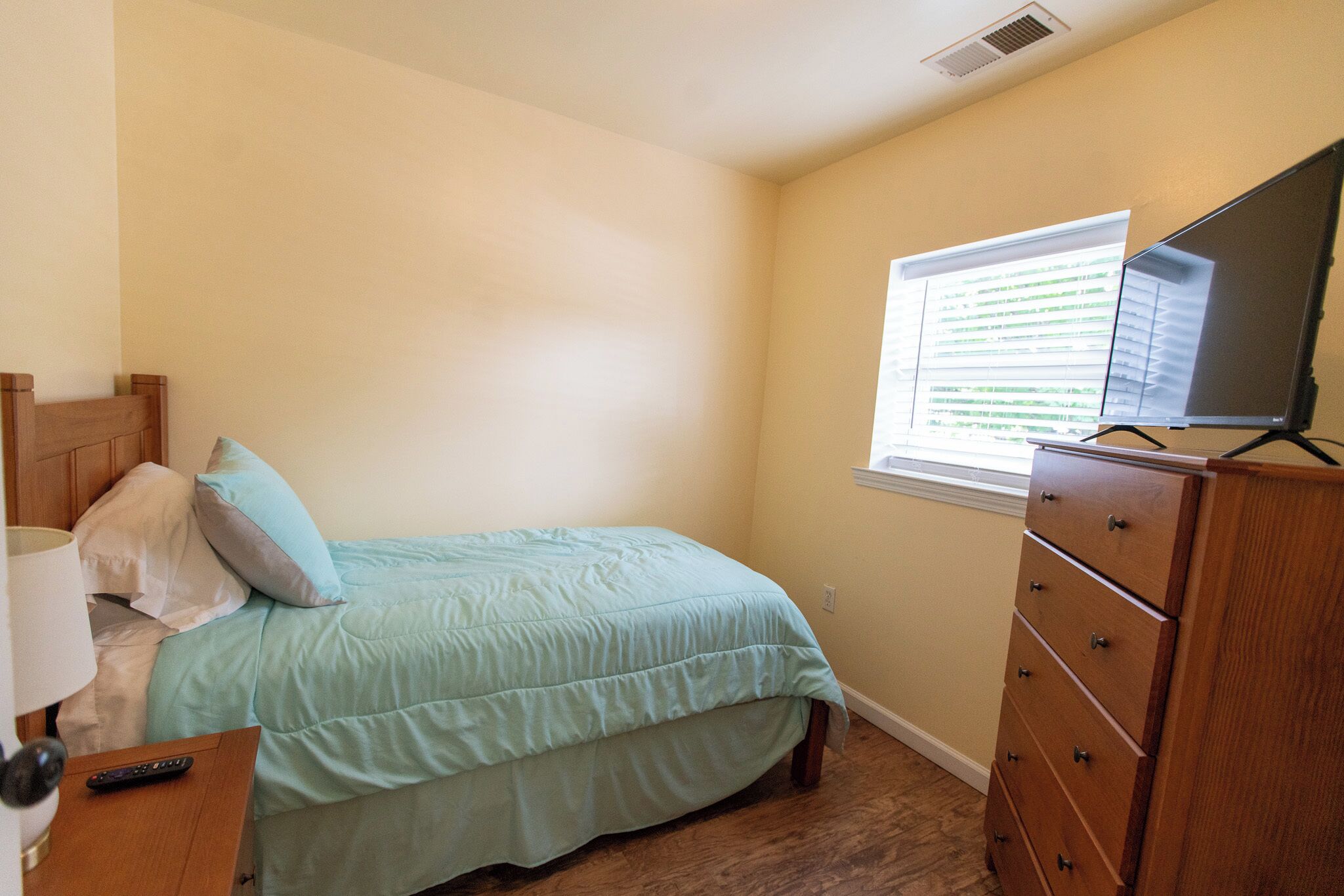 Un dormitorio con cama, cómoda, mesita de noche, lámpara, control remoto y ventana con persianas.