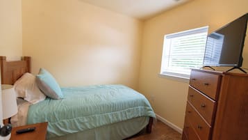 Un dormitorio con cama, cómoda, mesita de noche, lámpara, control remoto y ventana con persianas.
