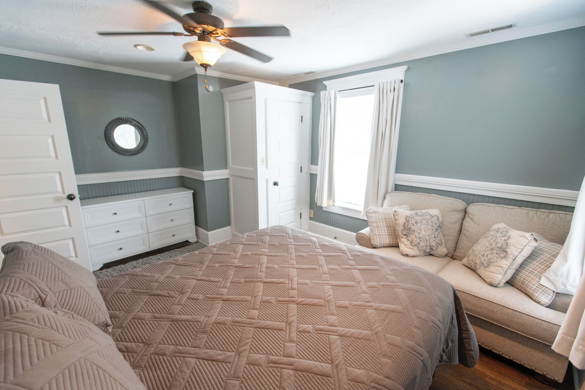 A bedroom with a large bed, a dresser, a ceiling fan, and a window with curtains.