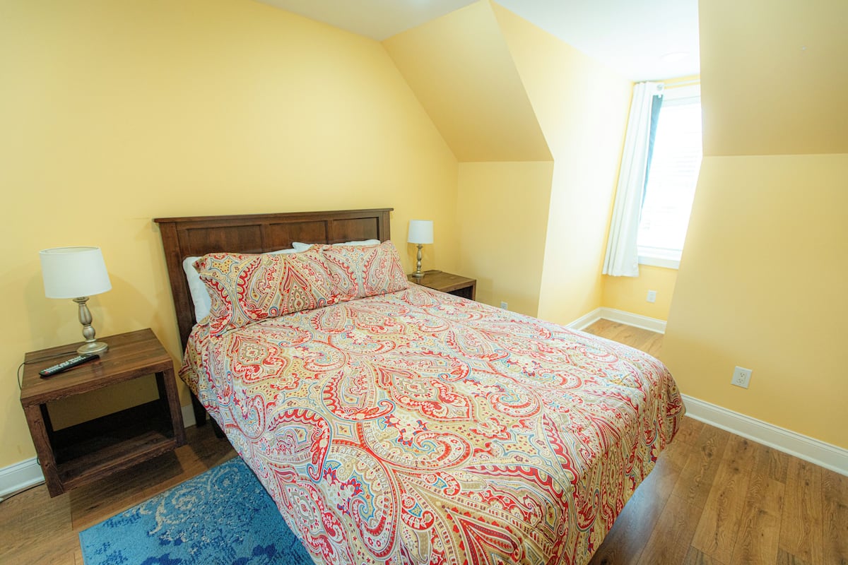A bedroom with a bed, two bedside tables, a lamp on each table, a blue rug, and a window with curtains.