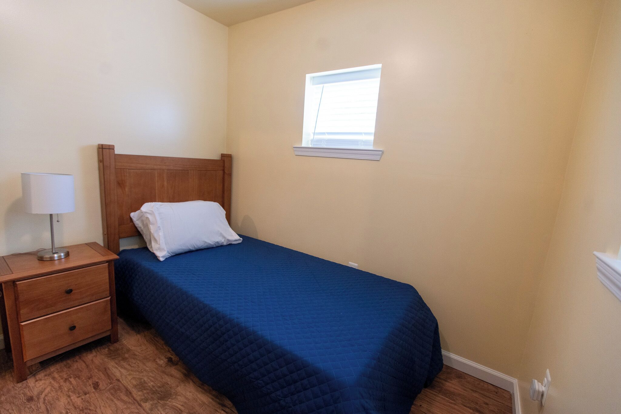 A bedroom with a single bed, a nightstand, a lamp, and a window.