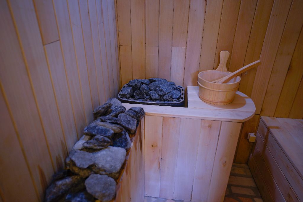 Un sauna avec des murs en bois, un mur en pierre, un plateau de pierres et un seau en bois avec une louche.