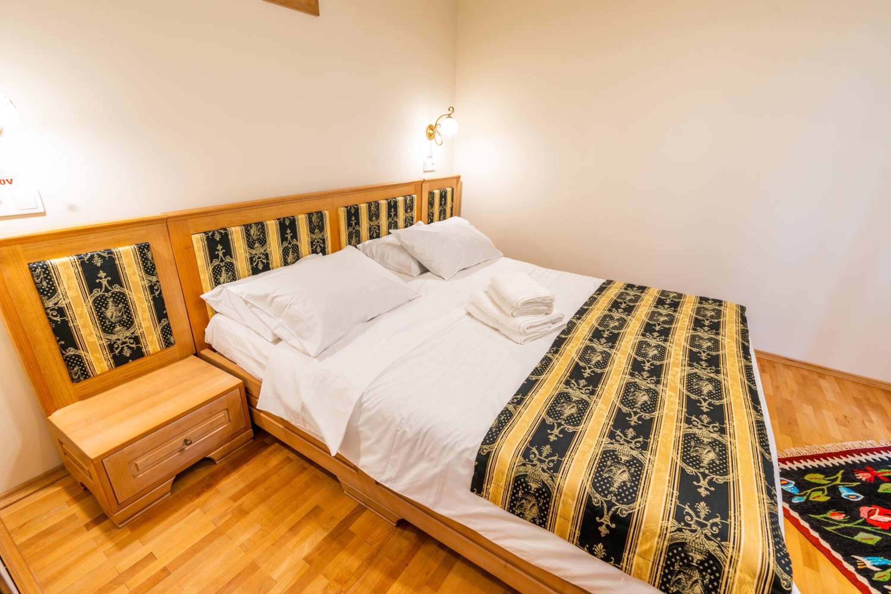 A neatly made bed with a patterned bedspread, a wooden nightstand with a drawer, and a wall-mounted light fixture.