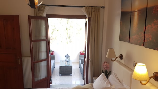 A hotel room with a bed, a lamp, a view of the outside, and a door leading to a balcony. - Santorini Family Apartments (Santorini)