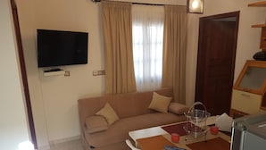 A living room with a beige sofa, a flat-screen TV mounted on the wall, a dining table with chairs, and a window with curtains. - Santorini Family Apartments (Santorini)