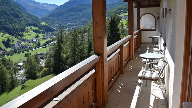 Ein Balkon mit Holzgeländer, einem kleinen Tisch und einem Stuhl. Der Balkon bietet Blick auf ein Tal mit Häusern und Bäumen.