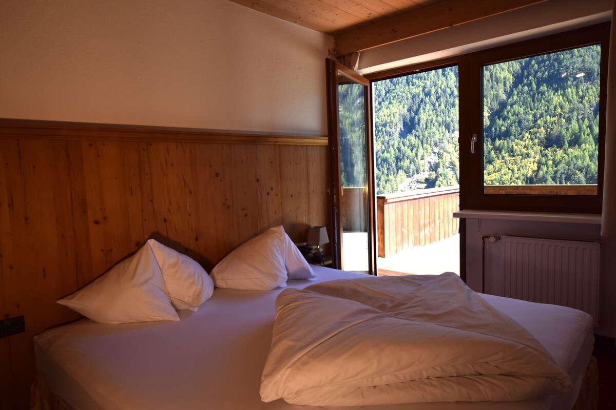 A bedroom with a wooden headboard, a large window with a view of a forest, and a bed with white bedding.