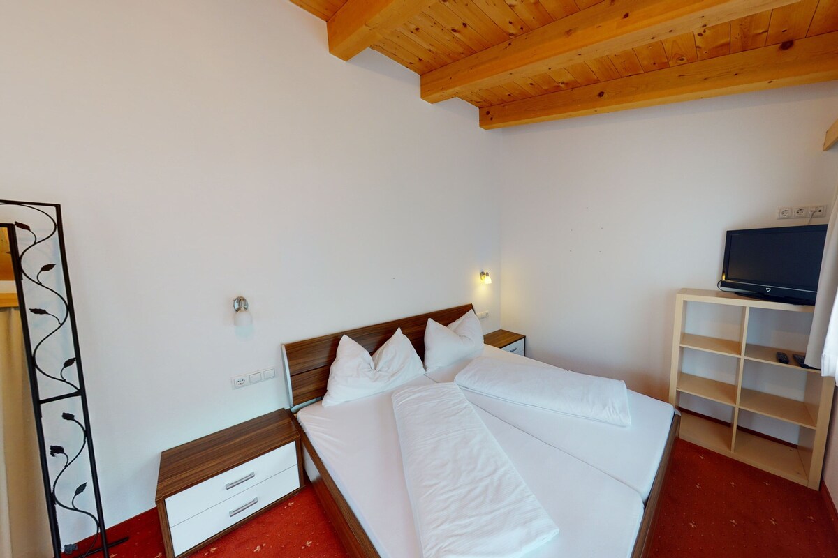 A hotel room with a wooden ceiling, a bed with white linens, a TV on a shelf, and a red carpet.