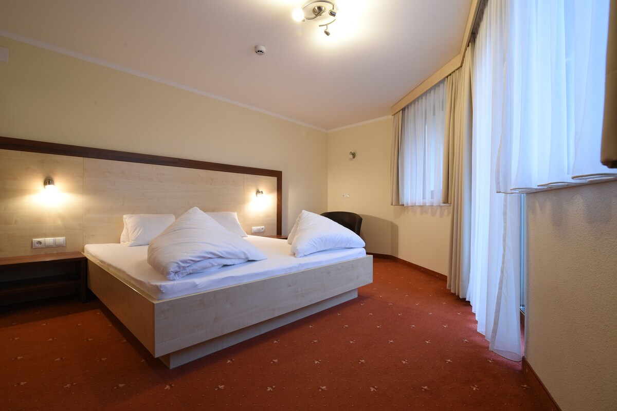 A hotel room with a large bed, a nightstand, a chair, and a window with curtains.