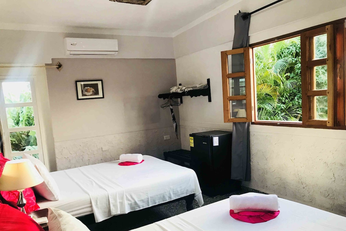 A room with two beds, a window with a view of greenery, a wall-mounted air conditioner, and a small black refrigerator.