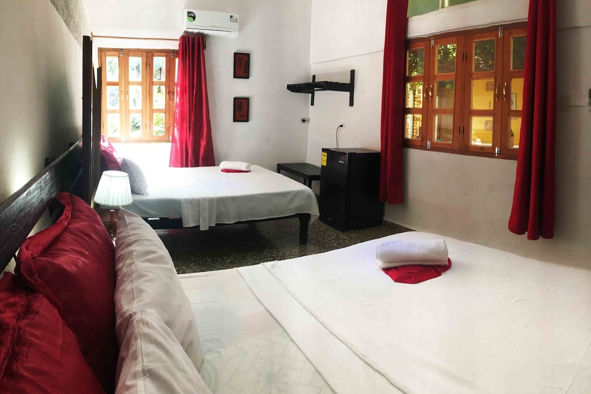A hotel room with two beds, a desk, a lamp, and a window with red curtains.