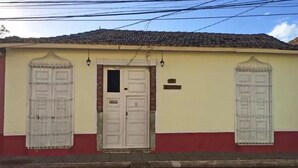 A building with a red and yellow facade, a white door, and two windows with metal grilles. - Casa Las Artes (Trinidad)