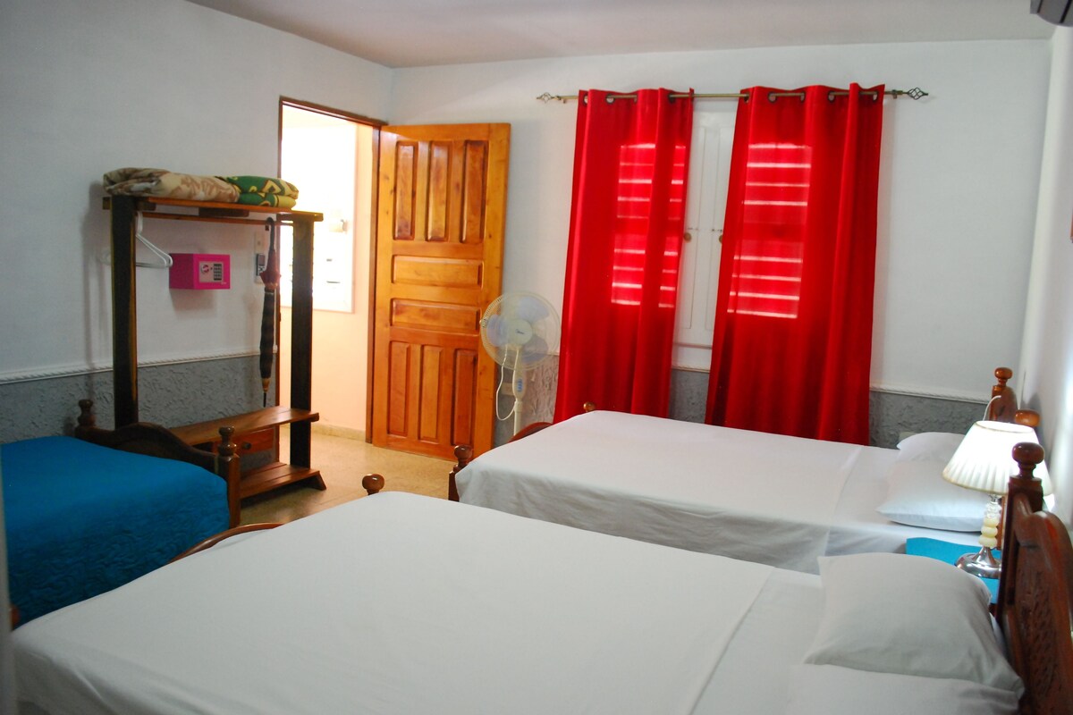 A bedroom with two beds, a wooden wardrobe, a desk with a chair, and a window with red curtains.