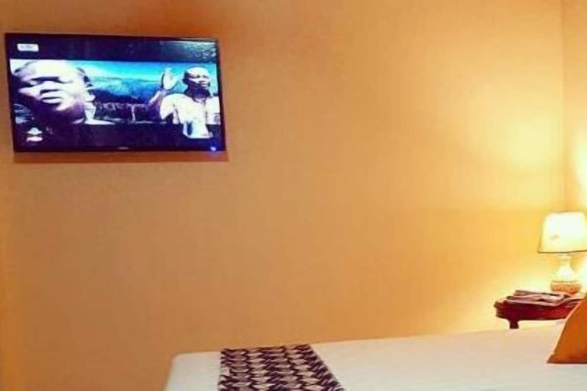 Quarto de hotel com cama, travesseiros, televisão na parede e mesa de cabeceira com abajur.