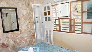 A bedroom with a stone wall, a bed with a blue and gold bedspread, a white door, and a window with a white blind. - Hostal Juan Fernandez (Trinidad)