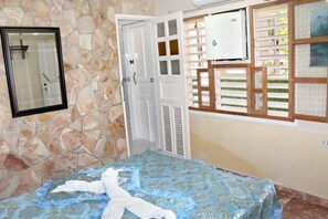 A bedroom with a stone wall, a bed with a blue and gold bedspread, a white door, and a window with a white blind. - Hostal Juan Fernandez (Trinidad)