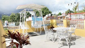 An outdoor patio with white metal chairs and tables, a covered seating area, and a view of the surrounding landscape. - Hostal Juan Fernandez (Trinidad)